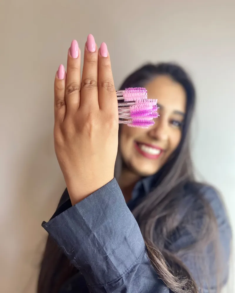 Mão da Grazzi com escovinhas de cílios, ela sorrindo ao fundo